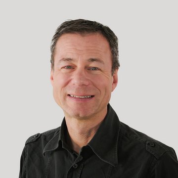Mann mit kurzem dunklem Haar und schwarzem Hemd vor hellem Hintergrund.