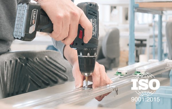 Eine Person bohrt an einem Werkstück; eingeblendet ist der Hinweis ISO 9001:2015.