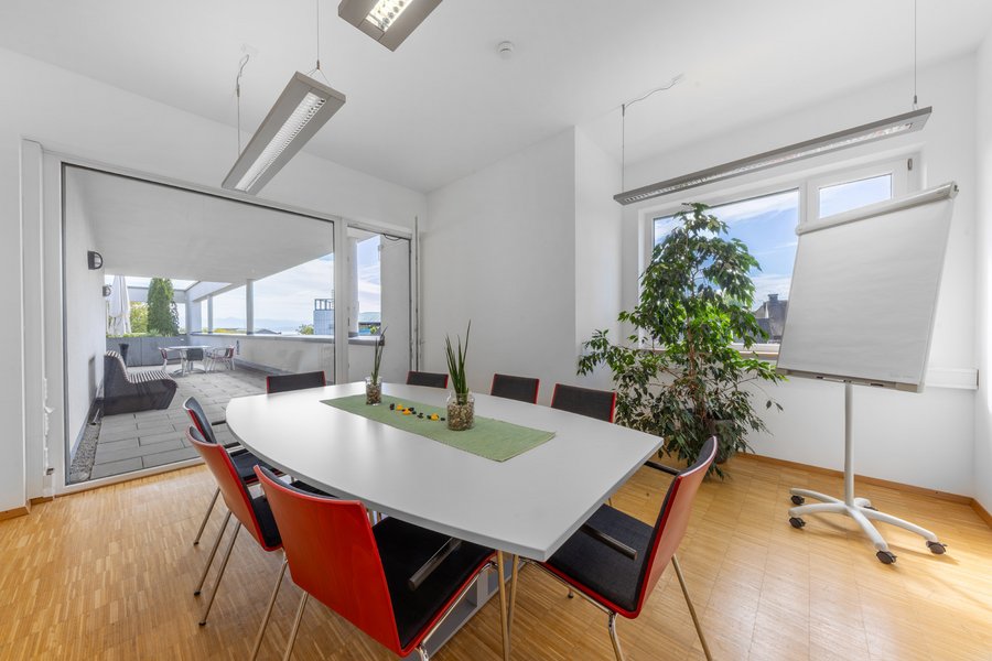 Heller Besprechungsraum mit Tisch, roten Stühlen und Fensterfront.