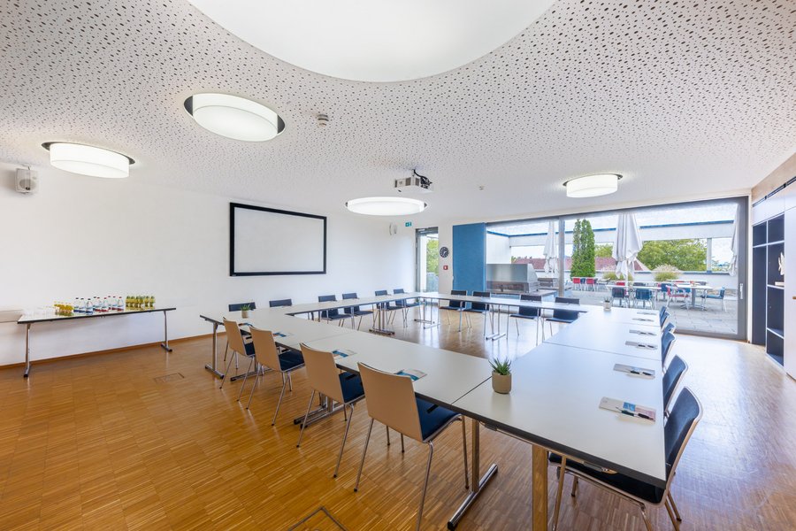 Großer Tagungsraum mit U-förmigen Tischen, Projektionsfläche und runden Deckenleuchten.