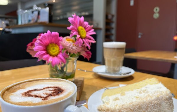 Eine Tasse Kaffee mit Blumen und einem Stück Kuchen auf einem Cafétisch.