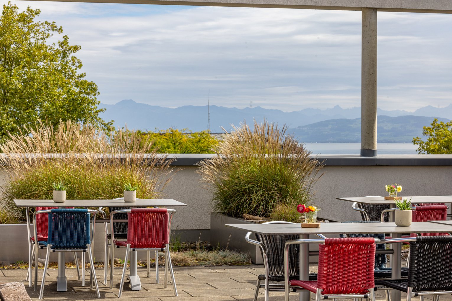 Dachterrasse mit roten Stühlen, Gräsern und Blick über Dächer zum See.