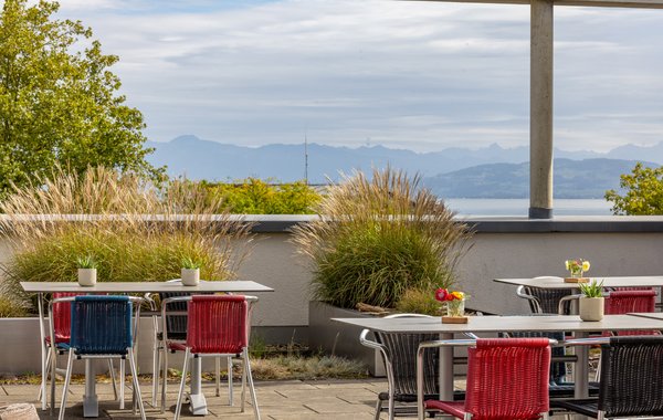 Dachterrasse mit roten Stühlen, Gräsern und Blick über Dächer zum See.