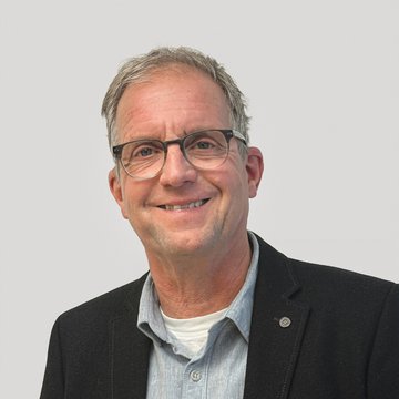 Mann mit Brille, Sakko und hellem Hemd vor hellem Hintergrund.