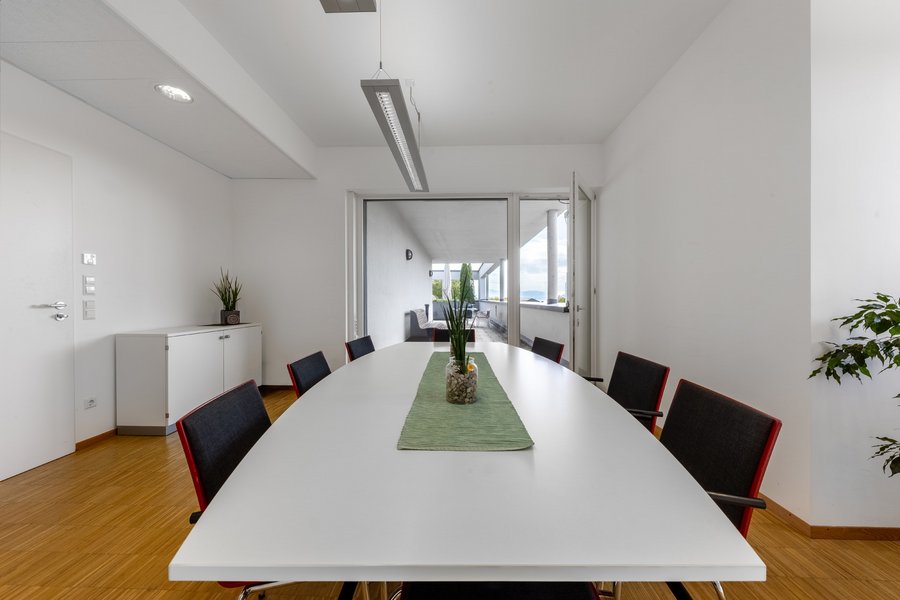 Heller Besprechungsraum mit langem Tisch, schwarzen Stühlen und Fensterfront.